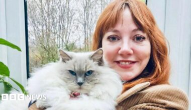 Diane Morgan, pictured holding a large, fluffy white cat which looks directly at the camera. Diane has long red hair and wears a brown corduroy jacket; she is smiling at the camera, standing in front of a window with trees in the background.