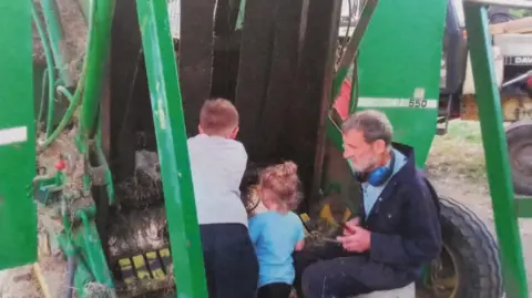 PA Media A man wearing navy overalls is sitting down at the base of a hay-baling machine he has two young children with him with their backs to the camera