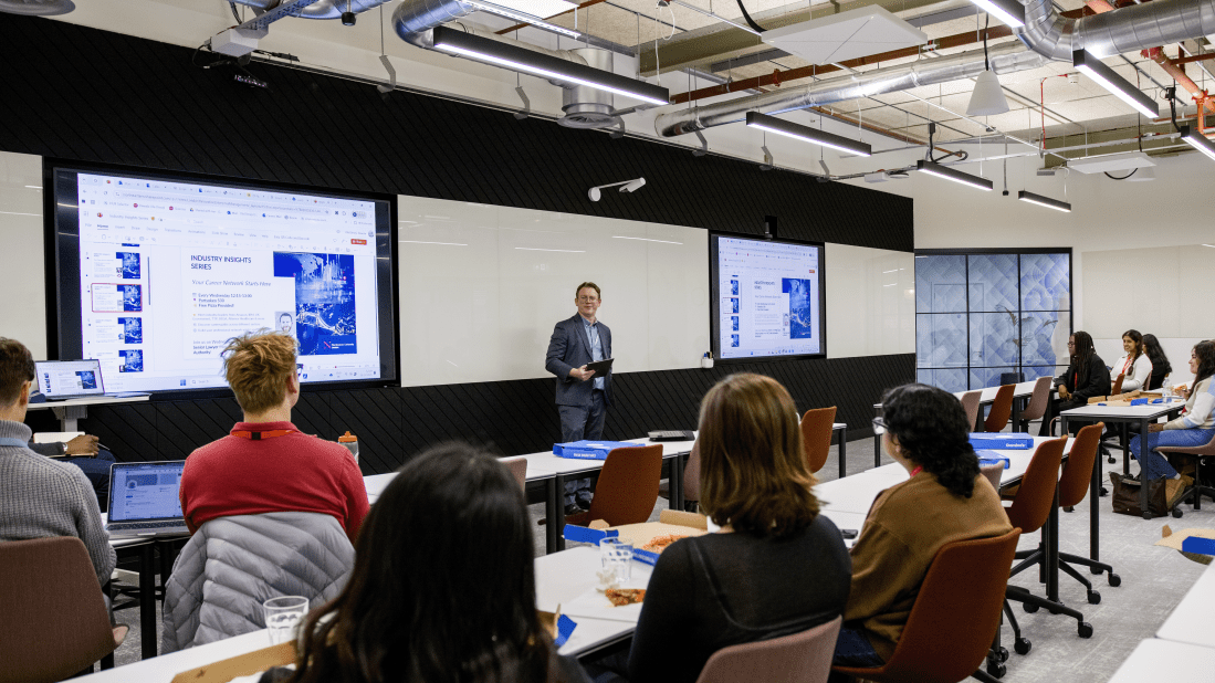 A suited man holding an electronic tablet device addresses a group of students