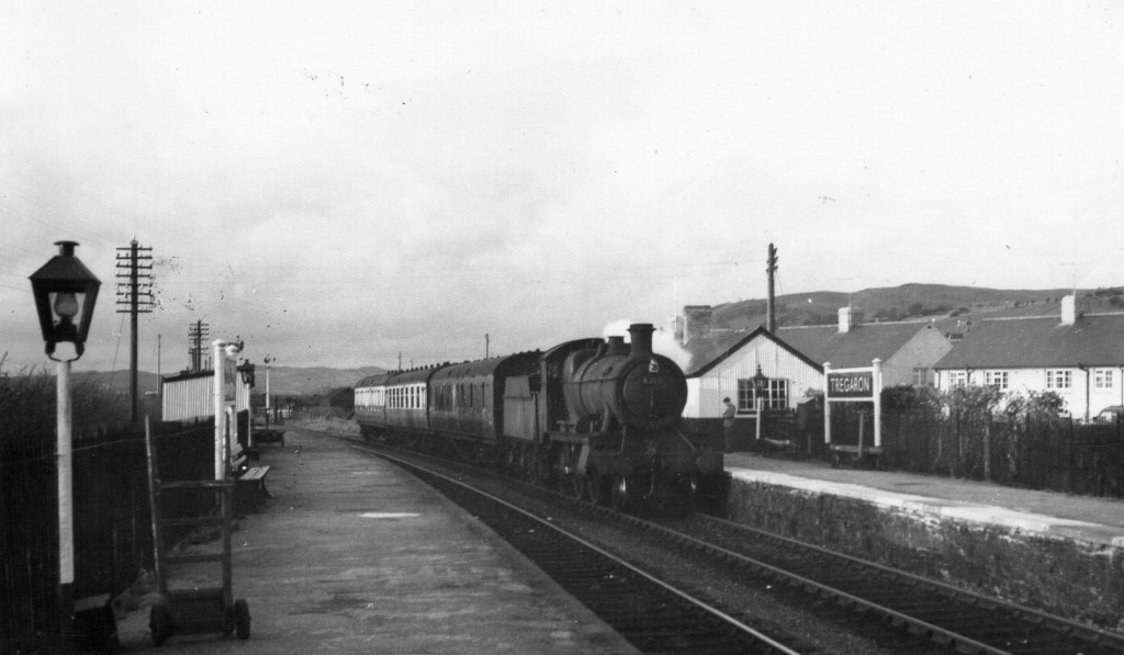 Black and white photograph showing a steam train arriving at Tregaron station, with platform furniture, station buildings, and surrounding homes visible. The image captures a moment from the mid-20th century, reflecting the region’s lost rail connectivity and the campaign to restore it.