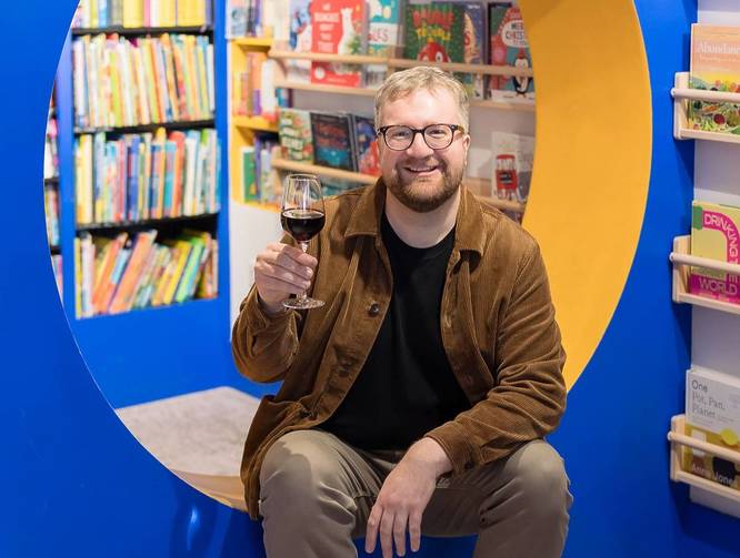 Owner of Backstory, Tom Rowley, holding up a glass of red wine at his store.