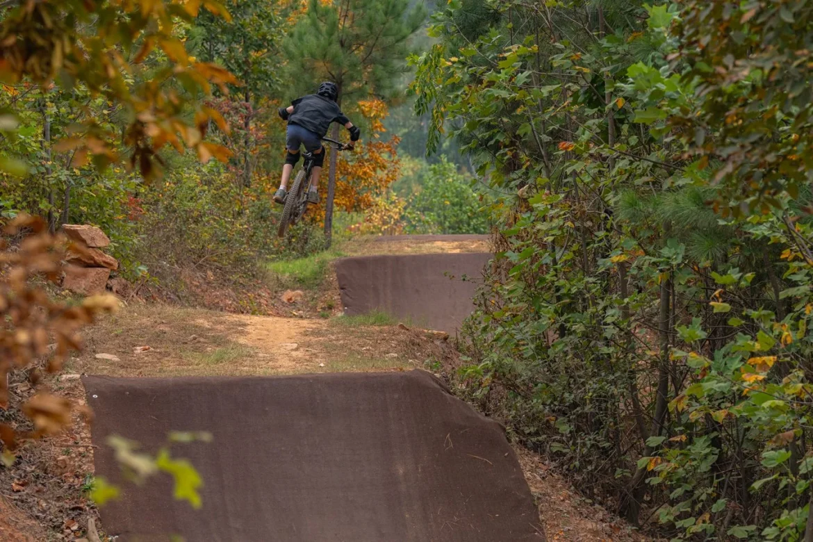 A mountain biker performing a jump over a dirt ramp in a forested area with autumn foliage.