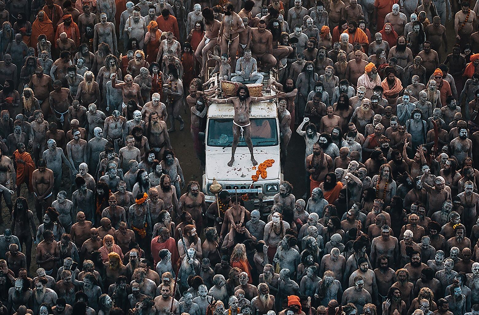 Aerial view of a large congregation of people covered in ash and paint, grouped around a vehicle, engaged in a vibrant spiritual gathering