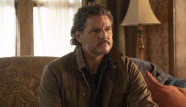 A bearded man in a brown jacket sitting on a sofa in a lounge.