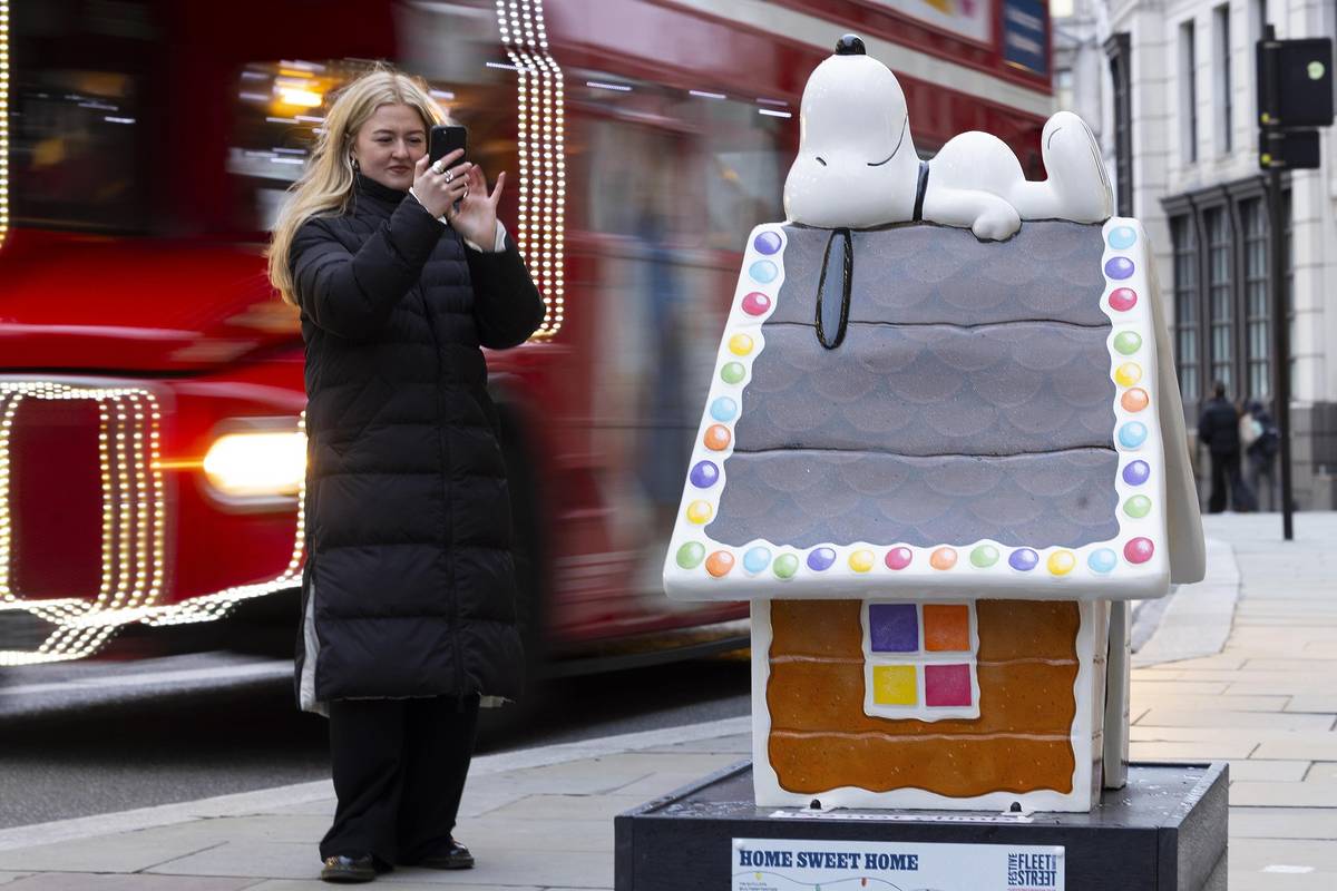 someone taking a photo of a sculpture of snoopy dozing on top of his doghouse as a bus goes past behind them