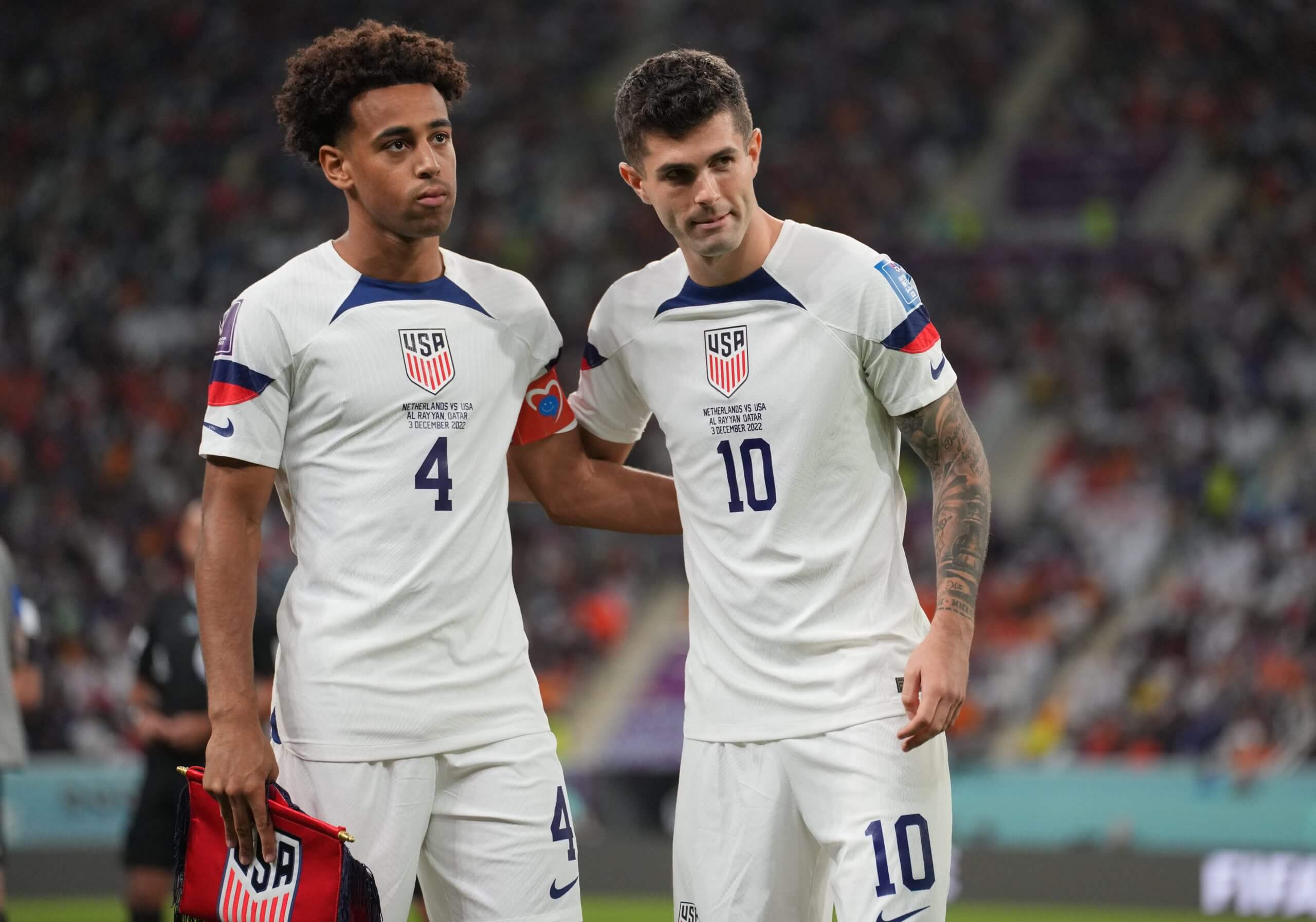 Tyler Adams, left, and Christian Pulisic, right, wearing their white USMNT uniforms, in a side embrace on the field with the crowd in the background.