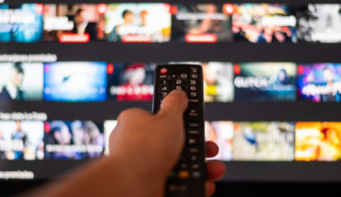 Hand Using A Television Remote Control in front of a TV with a streaming app on display and blurred in the background.