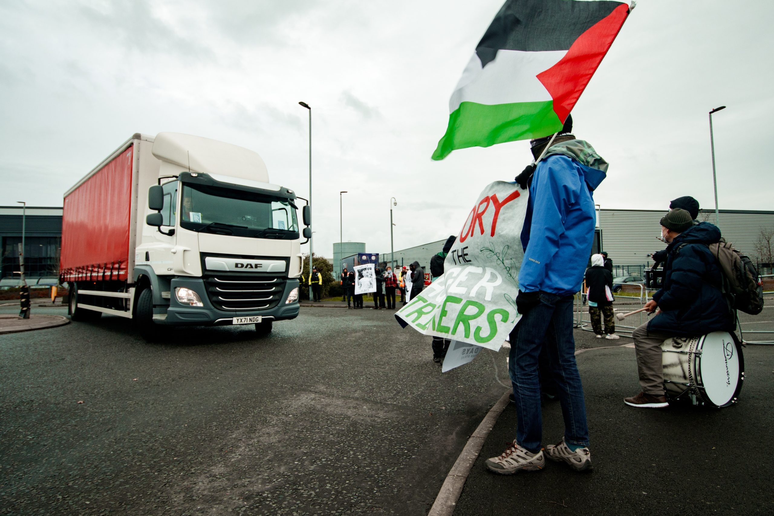 A delivery truck turns at a roundabout next to a row of protester holding flags and banners. 