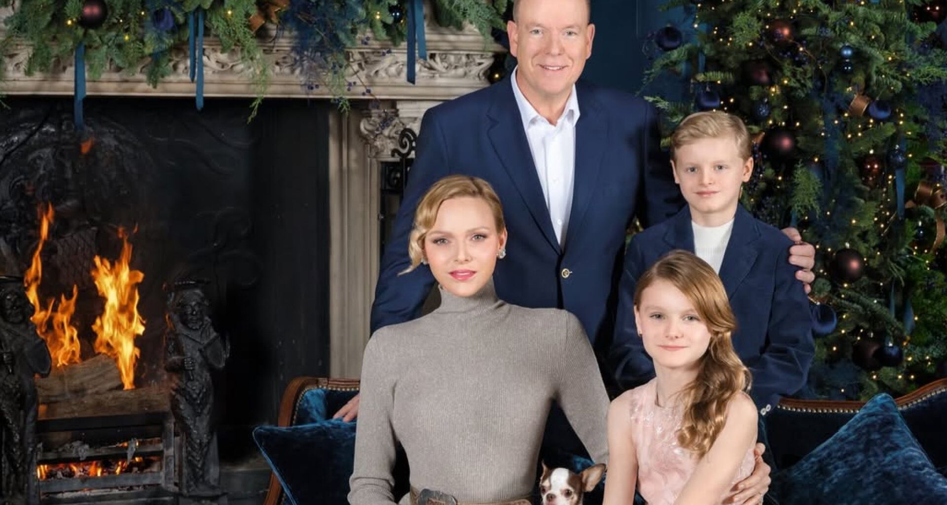 Princess Charlene wears a long taupe dress with a pleated skirt, long sleeves, and a turtle neck, and poses next to a burning fireplace and a Christmas tree with her pet dog, her twin children, Princess Gabriella and Prince Jacques, and husband Prince Albert