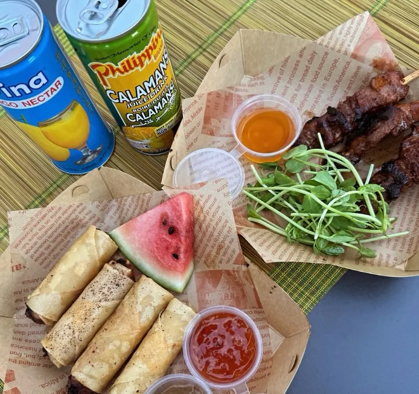 Takeout containers filled with lumpia and a slice of watermelon, and skewers of meat with pea shoots, next to cans of mango and calamansi juice.