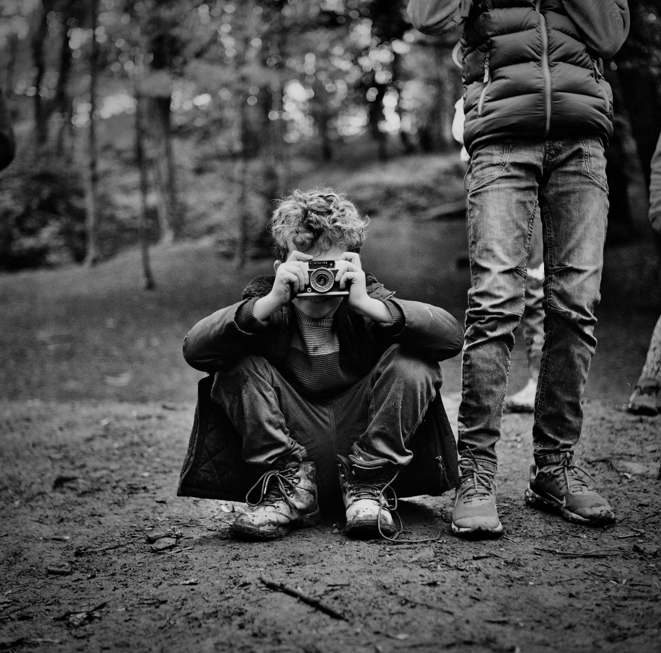 A boy squats in a park while holding a camera. 
