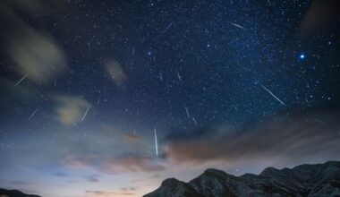 A series of white streaks of meteor showers are seen in a pale blue night sky over some mountain peaks at the bottom of the image