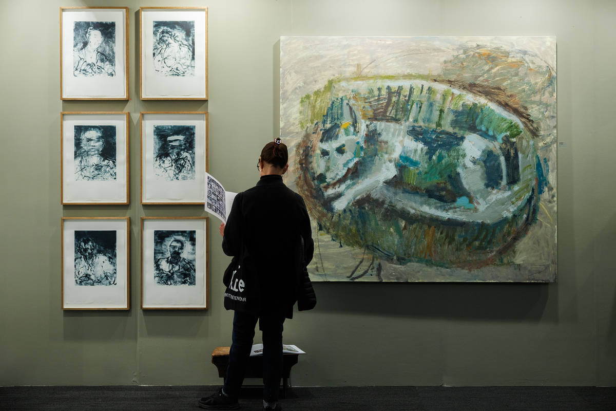 a visitor to the london art fair browsing a pamphlet while standing in front of six small-scale paintings arranged next to a larger human-sized painting