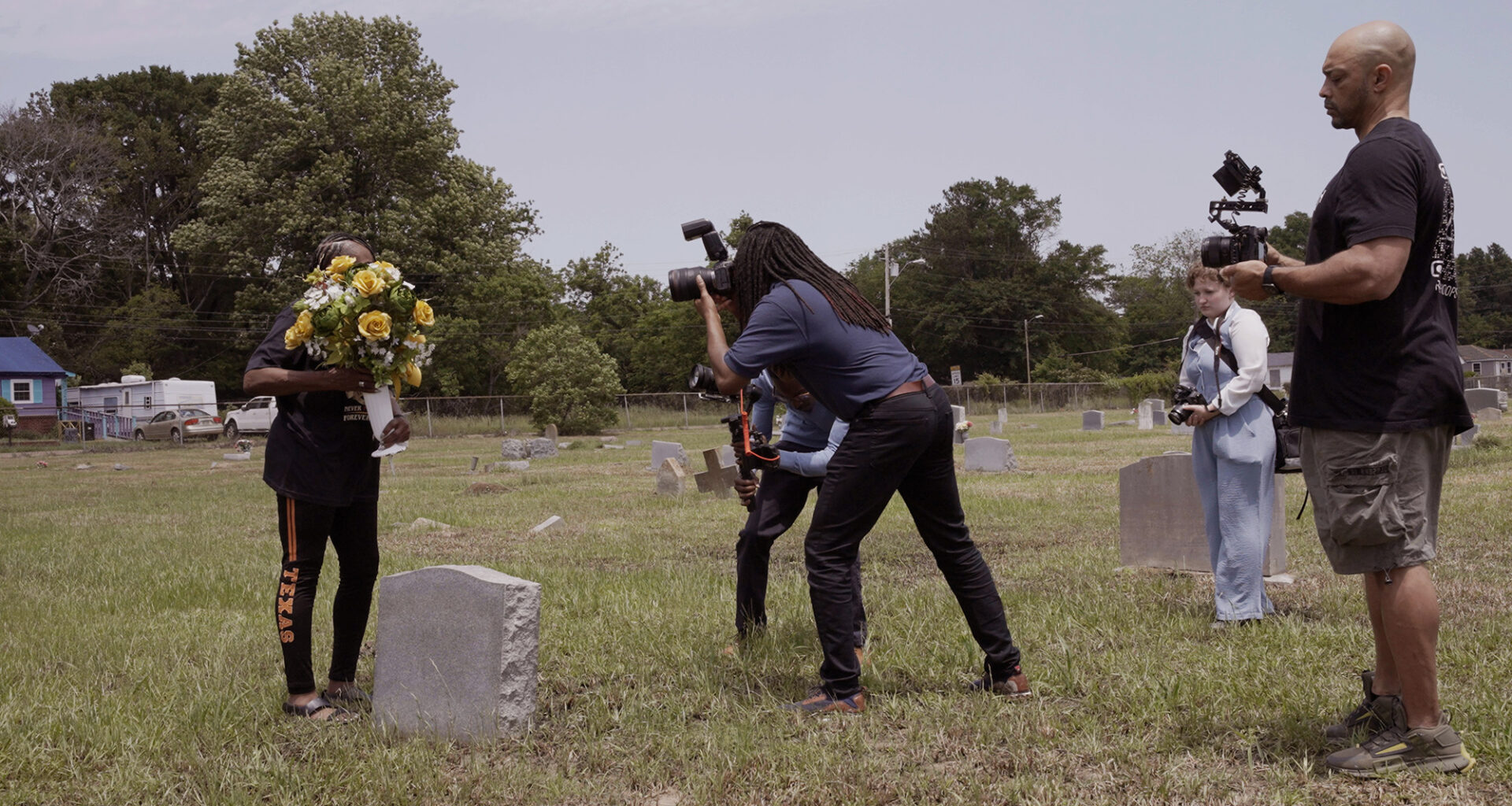 A film crew in a graveyard filming a person holding a bouquet of yellow flowers next to a tombstone