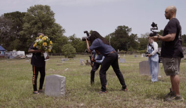 A film crew in a graveyard filming a person holding a bouquet of yellow flowers next to a tombstone