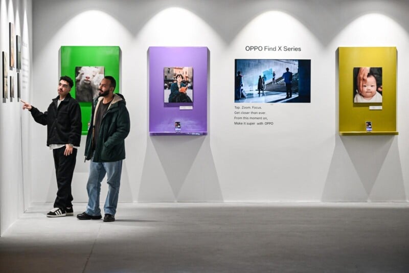 Two people view framed photos on a gallery wall; behind them, three vibrant displays showcase images and an OPPO Find X Series advertisement, emphasizing the phone's camera features. The gallery space is modern and well-lit.