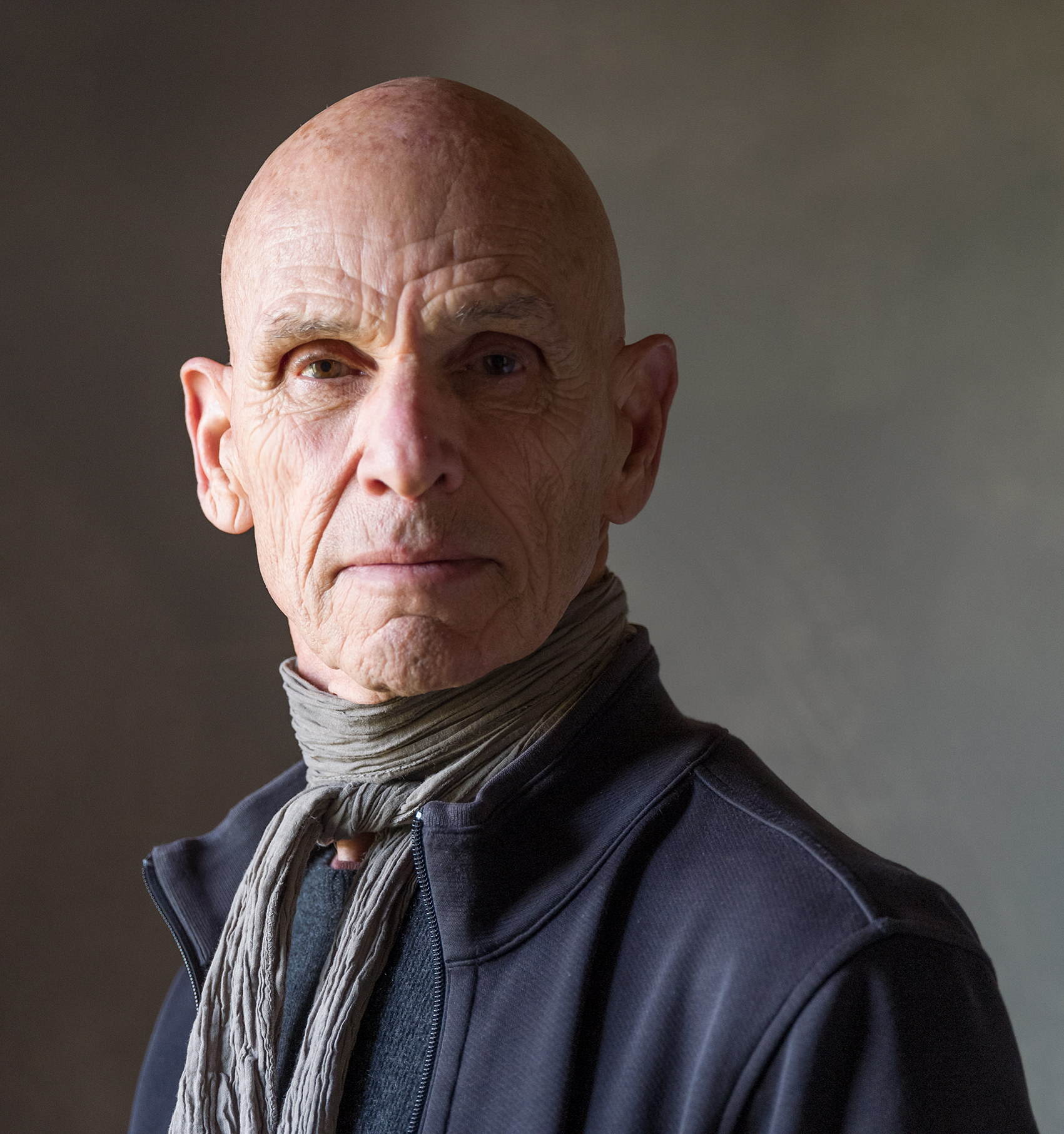 A portrait of Joel Meyerowitz, wearing a dark jacket and a textured scarf, against a softly lit background