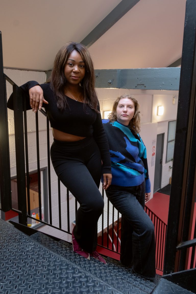 Two women lean against metal banisters in a stairwell