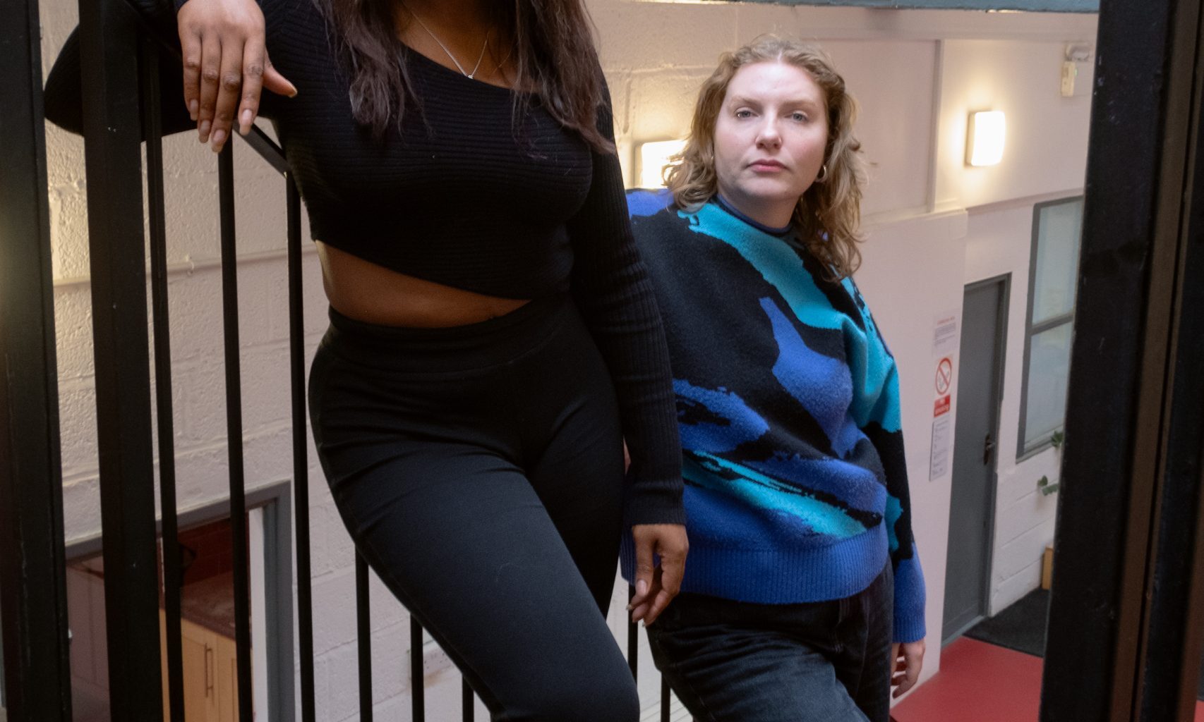 Two women lean against metal banisters in a stairwell