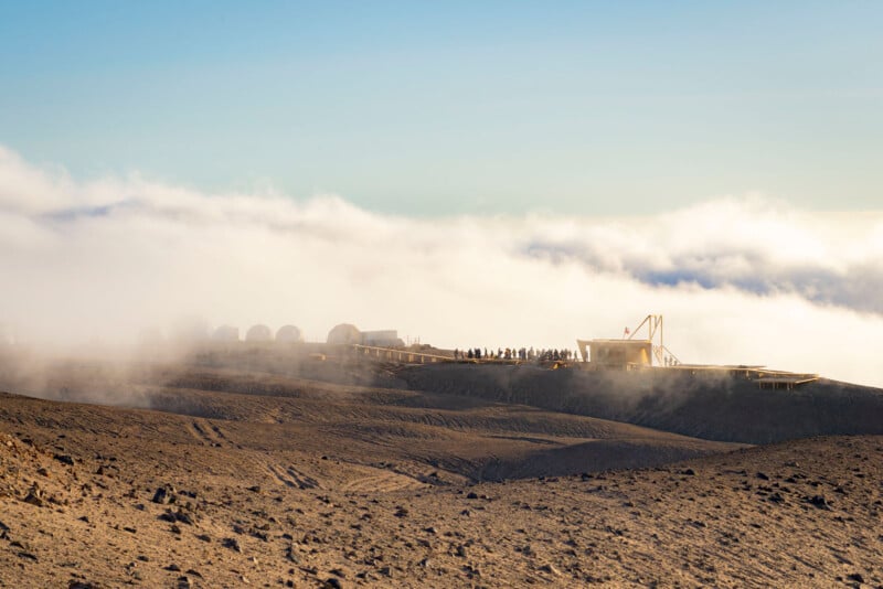 A modern observatory with multiple domes sits atop a barren, rocky mountain ridge, partially surrounded by mist and low clouds under a clear sky.