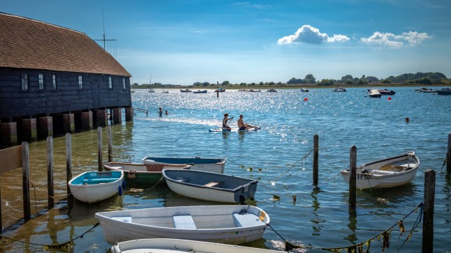 A scenic view of a couple on a paddleboard, floating next to the Bosham Sailing Club boathouse and static boats on the water in Bosham, West Sussex.
