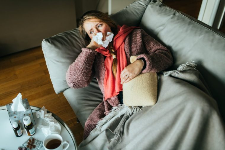 Woman lying on a sofa while having a flu and feeling sick.She using a heat pillow for her stomage and a bit of medicine standing on the table.
