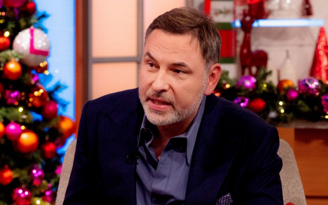 David Walliams on Lorraine. he's sitting on a chair wearing a dark jacket and shirt with a Christmas tree behind him.