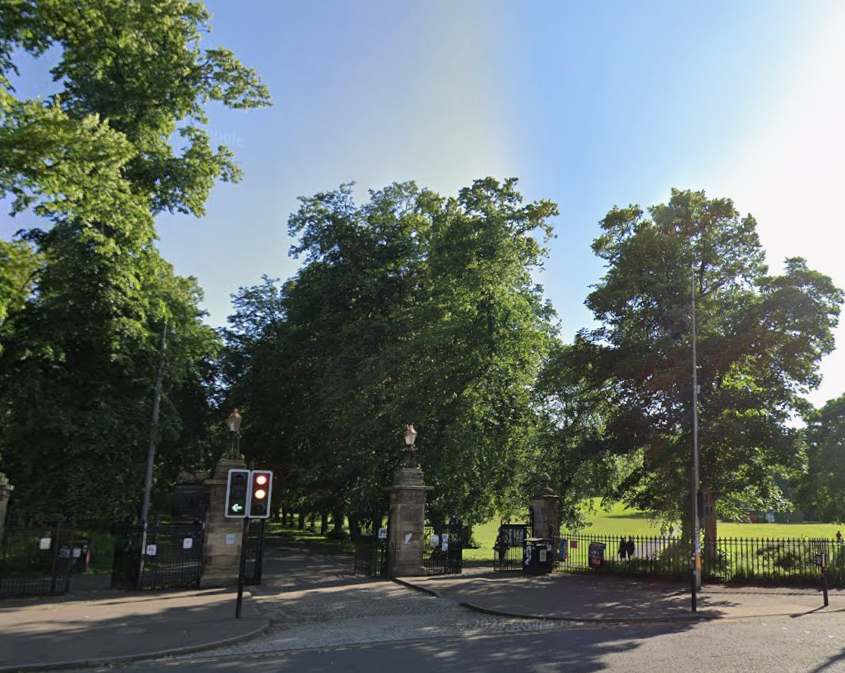 Entrance to a park with a wrought iron fence and stone pillars, with a traffic light showing red and a green arrow to the left.
