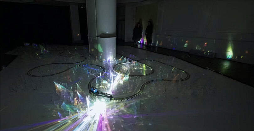 A dark room with a circular track surrounding a pillar, illuminated by colorful lights reflecting off transparent, crystal-like structures; two people stand in the background.