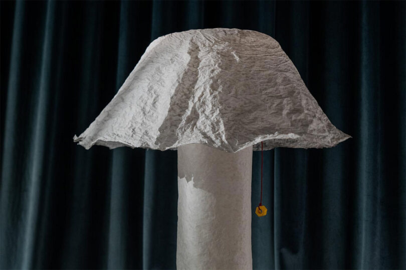 A floor lamp with a textured, crumpled white paper shade and a pull cord stands beside dark, pleated curtains, casting soft light.