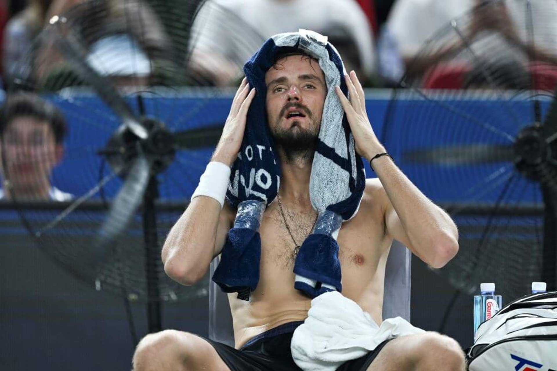 Tennis player Daniil Medvedev sits topless with a towel around his head, sweat dripping from his body.