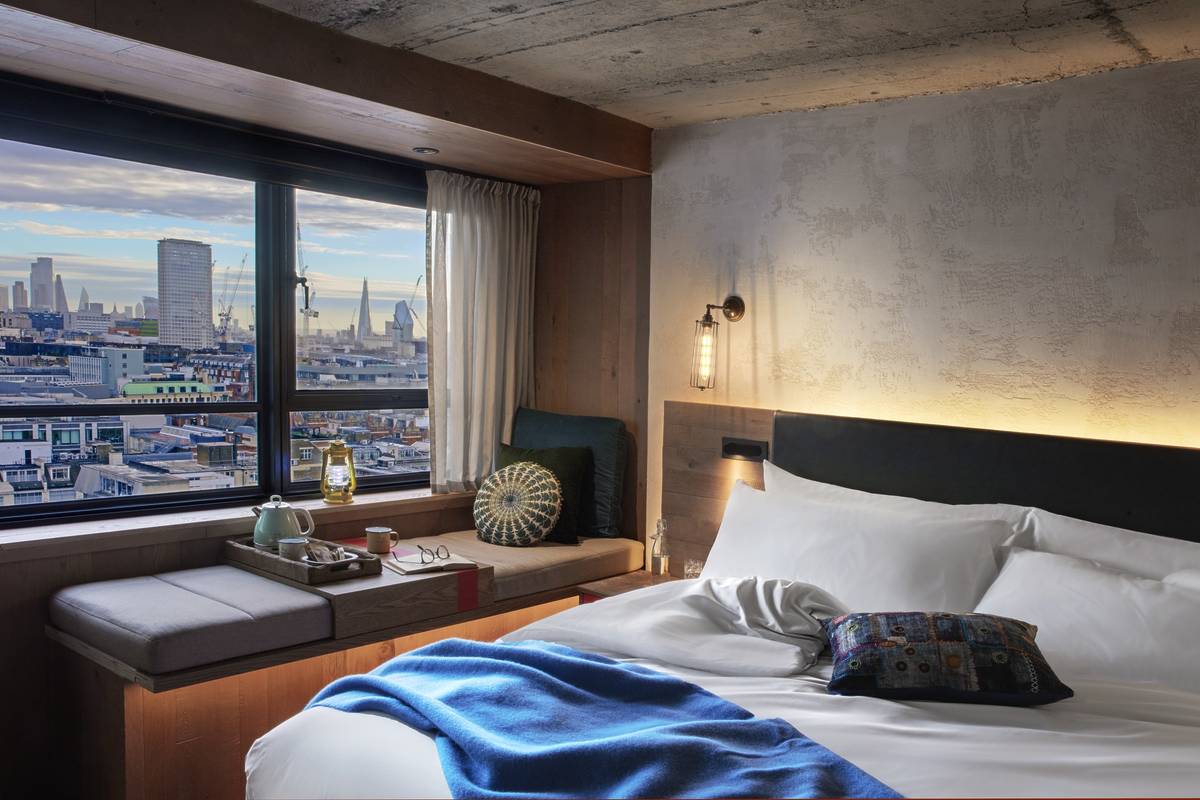 A bed and window seat in a hotel room at Treehouse, looking out onto a sweeping view of London