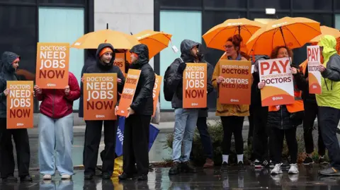 EPA Resident doctors hold orange signs saying 'doctors need jobs now' and 'pay restoration for doctors' while holding umbrellas outside Liverpool University Hospital on 14 November.