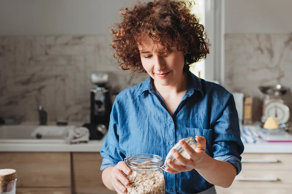 Person with curly hair in a casual shirt opens a jar in a kitchen, possibly preparing food. The setting suggests a focus on home or work-life balance