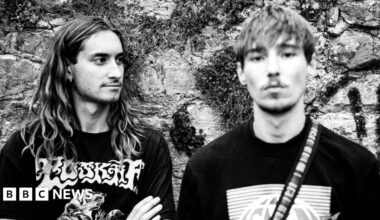 Two young men, one on the left with long hair and one on the right with shorter hair and a light beard, shot in black and white