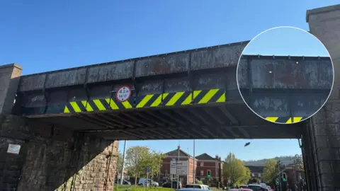 Graham Loveluck-Edwards A railway bridge over a road on which cars are queing. The bride has several small holes punched into it.