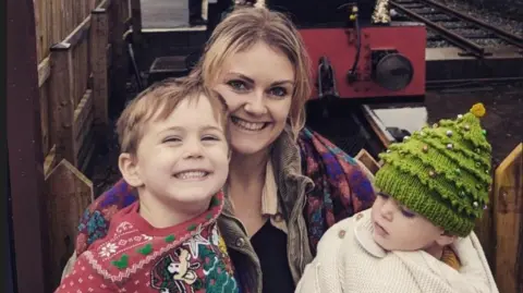 Mari Ellis Dunning A woman with long blonde hair is smiling while holding a young boy and a baby boy. They are sitting by a mini train. The older boy is smiling and wearing a red Disney-themed Christmas jumper and the baby has a Christmas tree-style woollen hat.