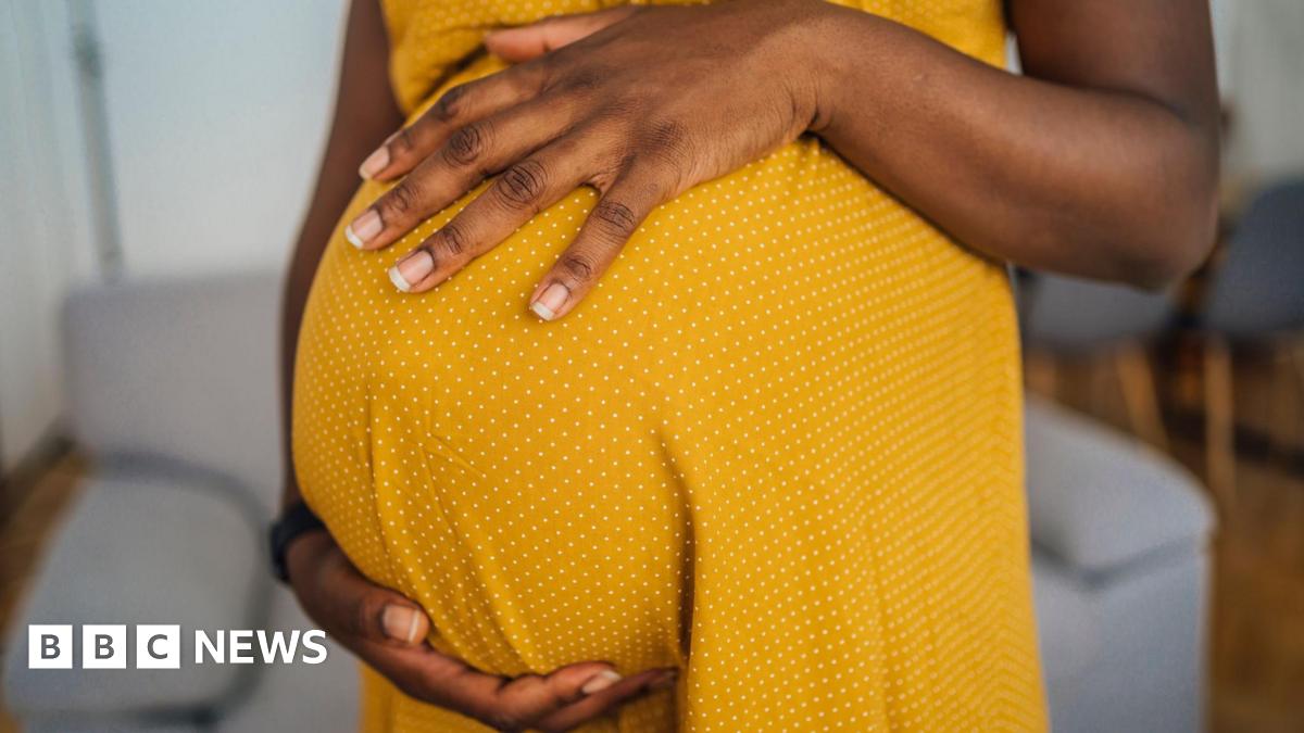 Close-up of pregnant black woman wearing a yellow dress with small white dots. The picture does not show the woman's face. She has both hands resting on her pregnant belly and she is standing in a room with a grey sofa behind her and some grey chairs and a table.
