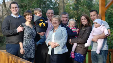 Rob and Dianne Parsons Photograph of Rob, Dianne and Ronnie (centre) with their two children, Lloyd and Kate, and their partners and three grandchildren. They are all stood out in a garden and trees can be seen behind. 