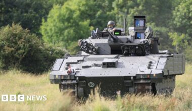 An AJAX armoured fighting vehicle, a large green tank, is seen being driven through a field.