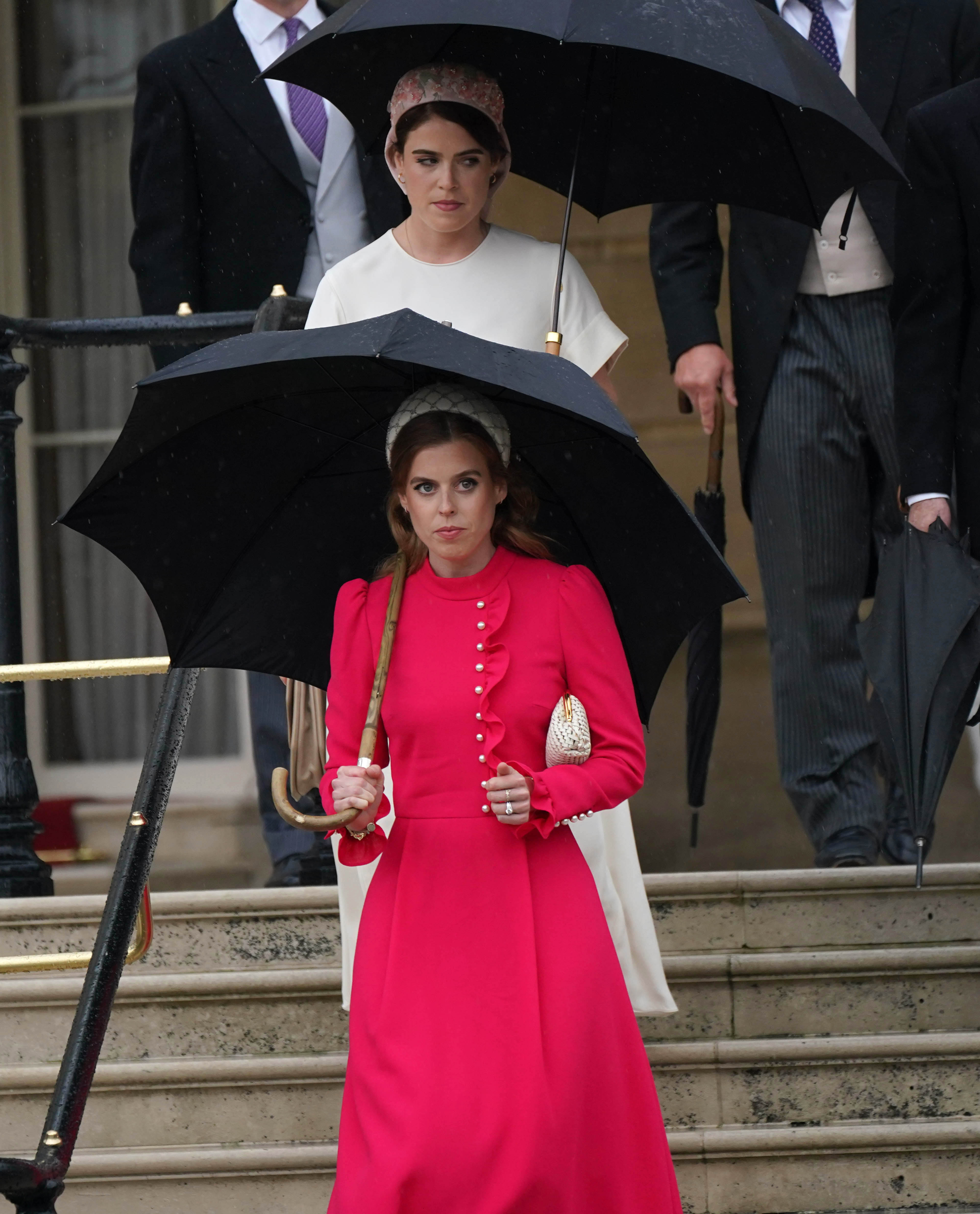 Princess Beatrice in a coral dress and Princess Eugenie in a white dress walking down a staircase and carrying umbrellas