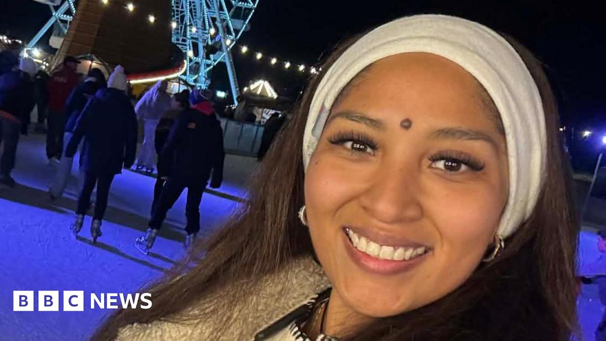 Joyce Leon Velasquez smiles at the camera on an ice rink at Christmas time. She has a white headband on and black coat. People can be seen ice skating behind her. There is also a large Ferris wheel that is lit up.