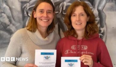 Two women, one in a red hoodie and one in a cream sweater, pose together smiling and holding up a white certificate for the camera