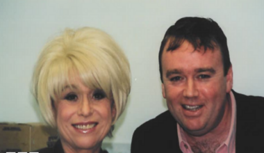 A blonde haired woman in a black top stands next to a man in a black blazer and pink shirt smiling at the camera.