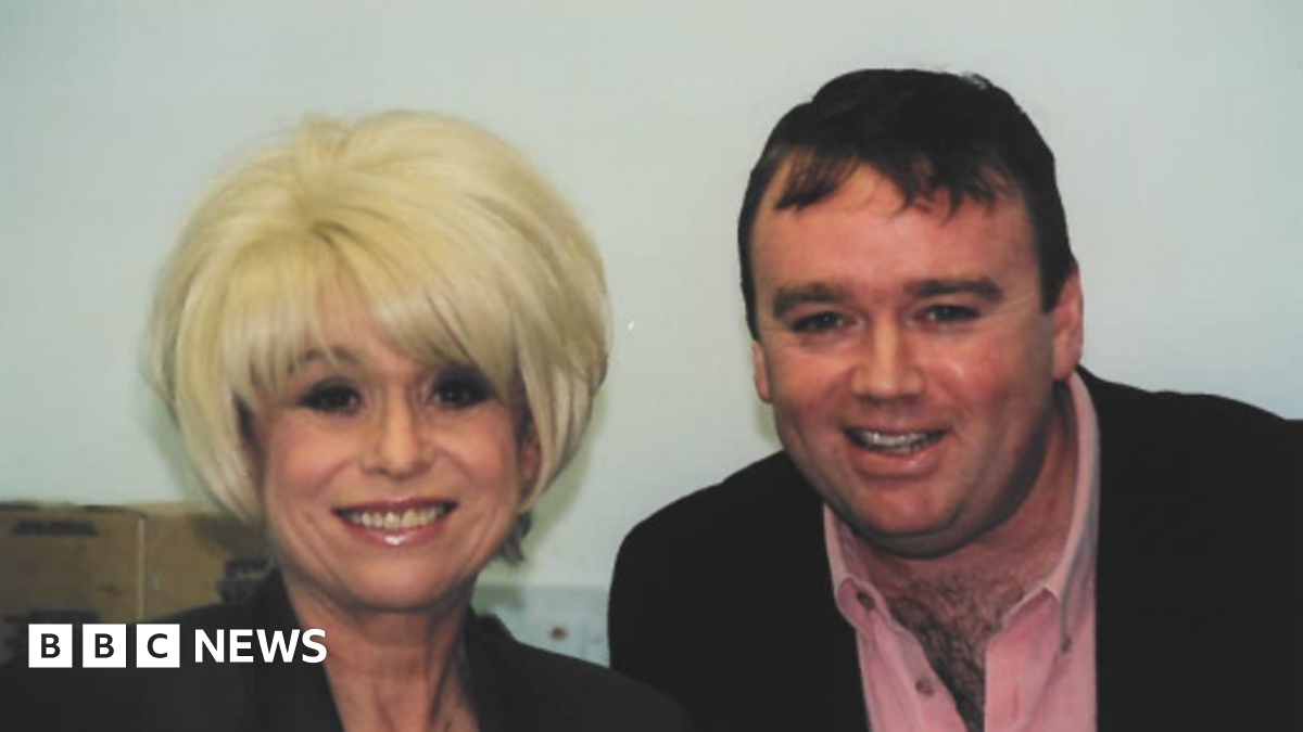 A blonde haired woman in a black top stands next to a man in a black blazer and pink shirt smiling at the camera.