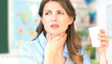 A woman with long brown hair and wearing a blue blouse clutches her throat with her hand, looking pained, while holding a white cup of something to drink