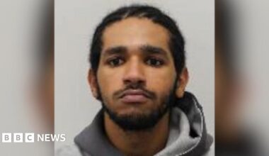 A young man with dark curly brown hair standing by the side of a road. He is wearing a black t-shirt and behind him are yellow crops and blue skies.