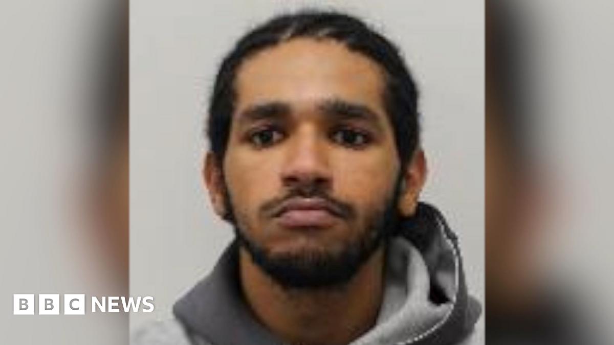 A young man with dark curly brown hair standing by the side of a road. He is wearing a black t-shirt and behind him are yellow crops and blue skies.