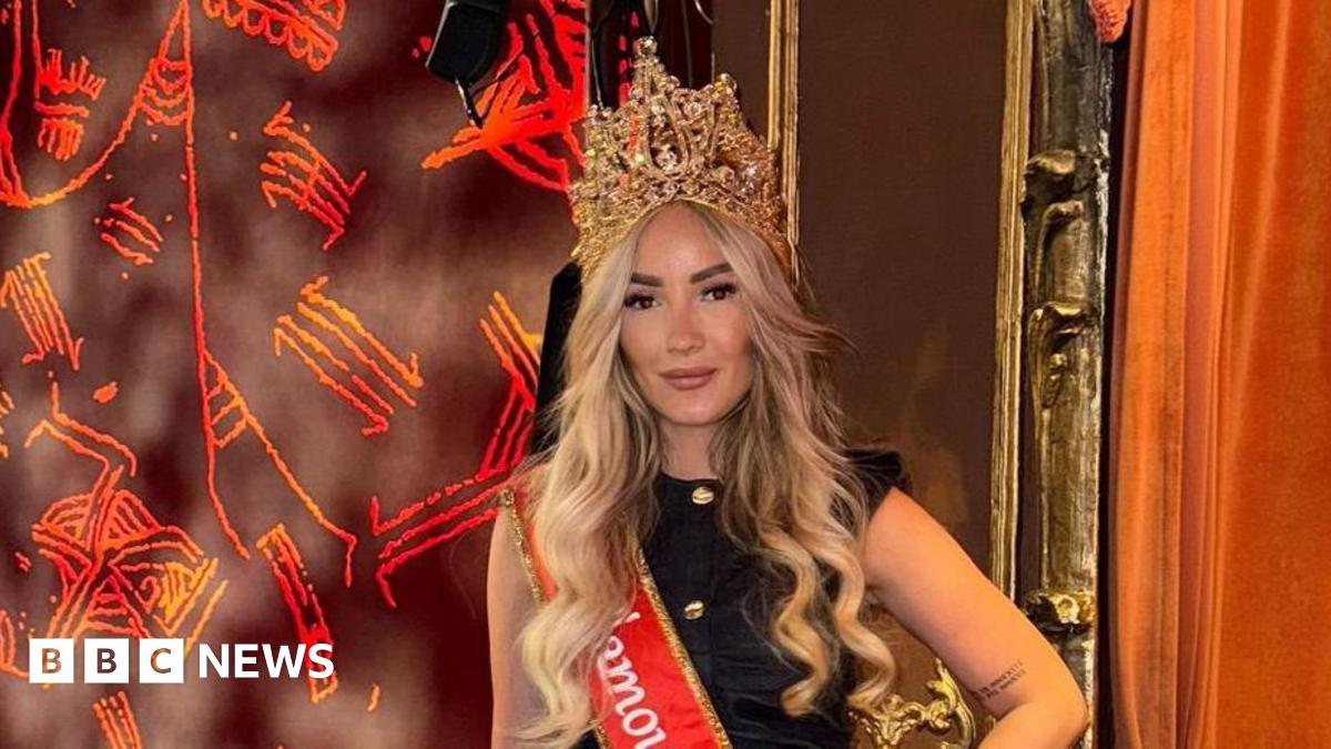 The image shows a blonde woman standing in a theatre box, wearing a crown and a bright red sash that reads "Ms Glamour UK 2025".