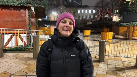A woman in a black puffer coat and a pink wooly hat is smiling at the camera and standing in front of a closed Christmas market